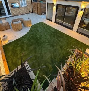 Patio Design with fake grass and entertainment area and warm lighting by Green Capital Landscaping and Gardening side view